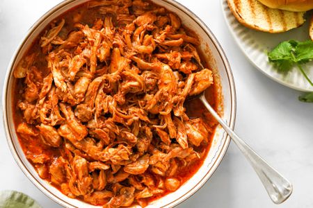 Bowl of Pulled Chicken With a Spoon Next to a Plate With a Toasted Bun and Spinach and a Light Green Table Napkin