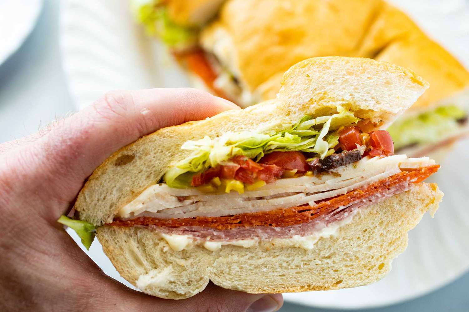 A hand holding a slice of a picnic sandwich layered with meat, cheese and toppings.