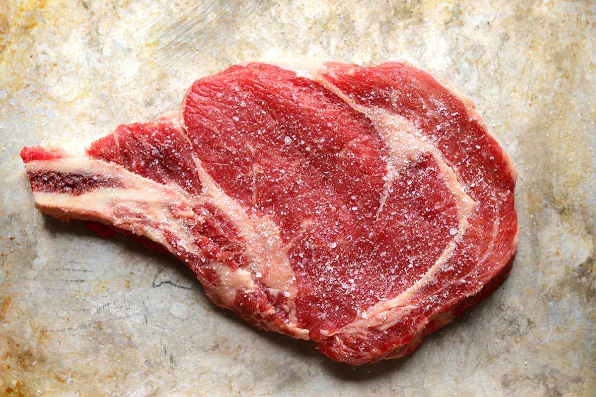 A bone-in raw steak seasoned with salt and set on a worn baking sheet.