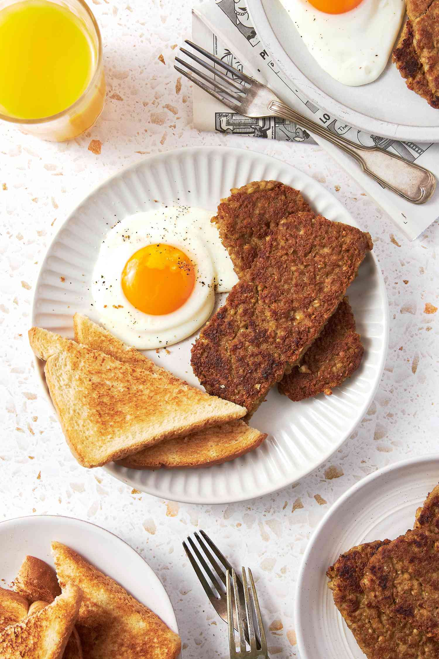 Plate With Sliced Goetta, a Sunny Side Egg, and Toast, and in the Surroundings, a Glass of Orange Juice, Another Plate With a Serving of Breakfast, a Plate With Sliced Goetta, a Plate of Toast, and Forks on the Counter