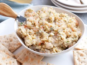 Bowl of Shrimp Butter with a Cheese Knife, Sitting on a Platter of Crackers at a Table Setting with a Glass of Wine, Stack of Plates, and a Kitchen Linen