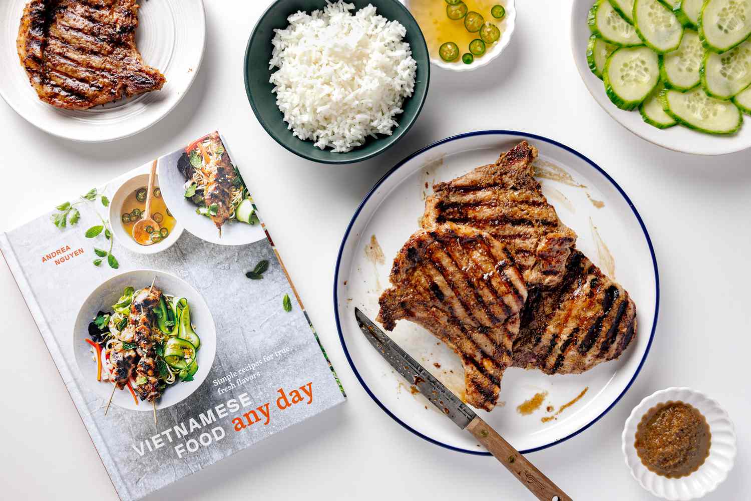 table spread of food with Andrea Nyugen's lemongrass pork chops on a serving plate, a pork chop on a smaller plate, a bowl with sliced cucumbers, a small bowl with sauce, a bowl of rice, and Andrea Nyugen's cookbook "Vietnamese Food Any Day"