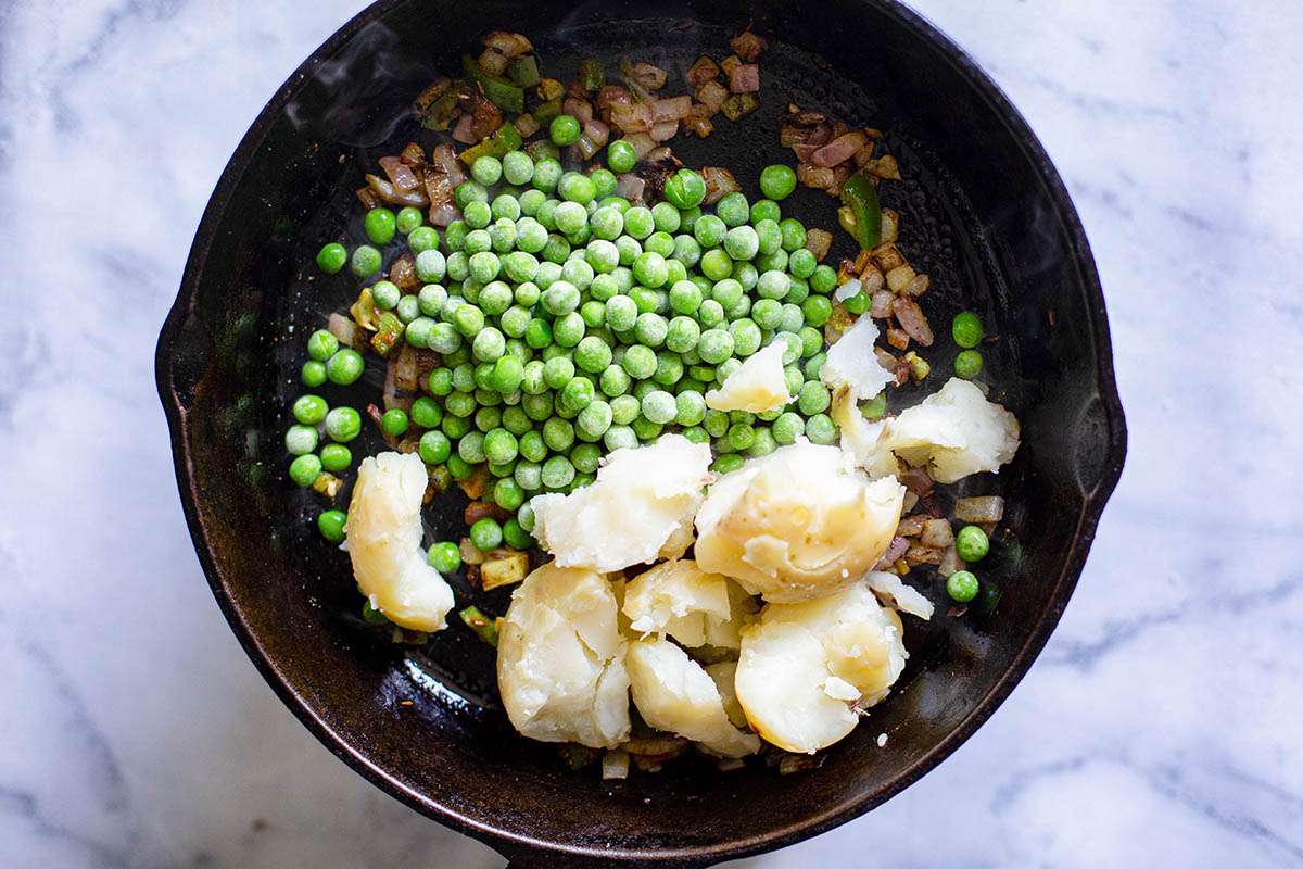 Onions, spices, peas, potatoes and jalapenos being toasted in a black skillet for vegetable samosa filling.