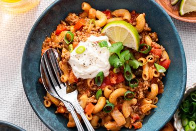 Bowl of One-Pot Taco Pasta Topped With Sour Cream, Sliced Scallions, a Sprig of Cilantro, and a Lime Wedge, and in the Surroundings, a Bowl With Lime Wedges, a Bowl With Sliced Green Onions, and a Glass With a Drink