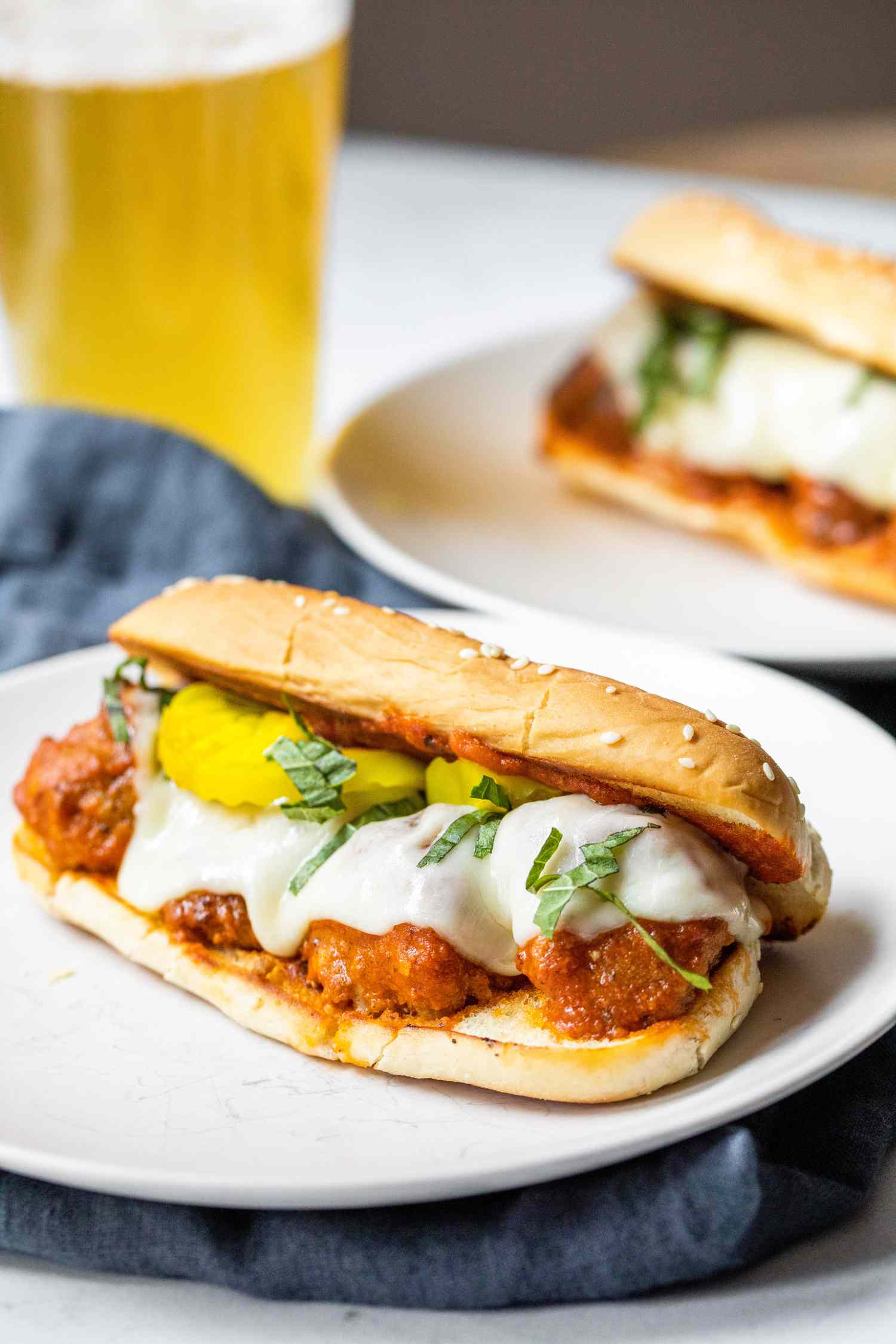 A Plate of Meatball Sub Topped with Banana Peppers and Basil, and in the Background, a Plate With Another Sub and a Glass of Beer