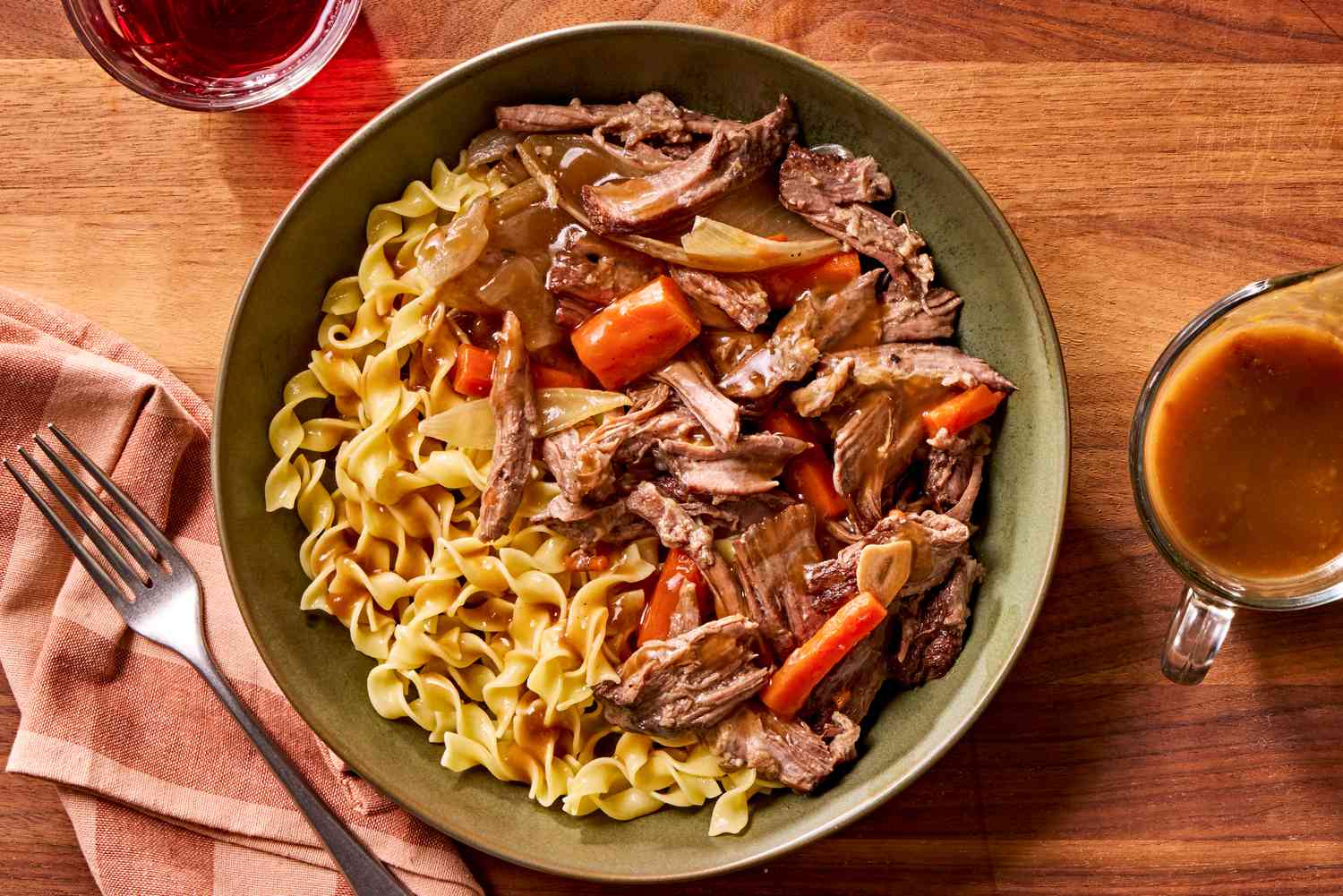 Overhead view of a plate of pot roast with carrots, onions and noodles next to a form on a napkin, a cup of gravy and a drinking glass