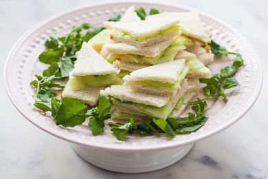 Triangle cut cucumber tea sandwiches are stacked on a cake stand and encircled with fresh herbs. Sliced cucucumbers are visible inside each one.
