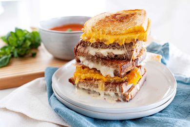 Grilled cheese sandwiches cut in half and stacked ontop of each other. Alternating with orange and white cheese with a bowl of tomato soup in the background.