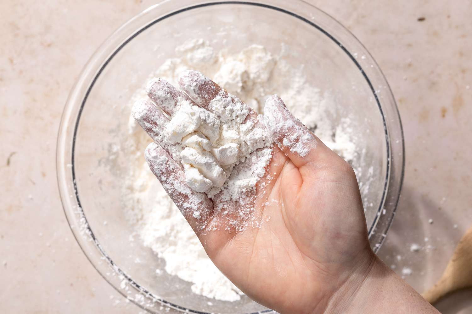 Overhead view of a clear glass bowl of the dry ingredients with a hand holding chunks of butter and flour mixed over the bowl for Mashed Potato Biscuits recipe