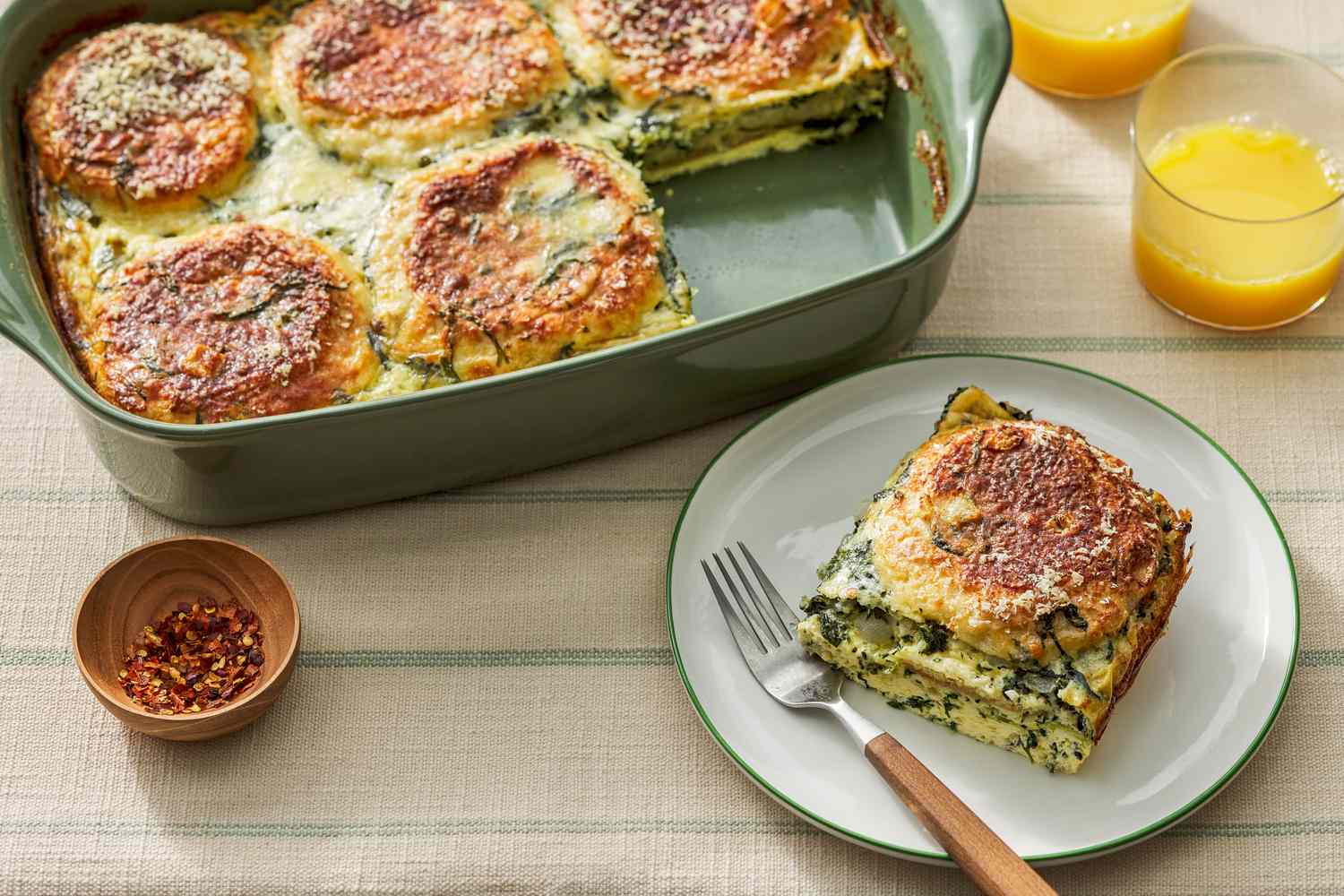 Serving of spinach florentine breakfast casserole on a plate, and in the surroundings, a casserole dish with more, a small bowl with crushed pepper, and glasses of orange juice