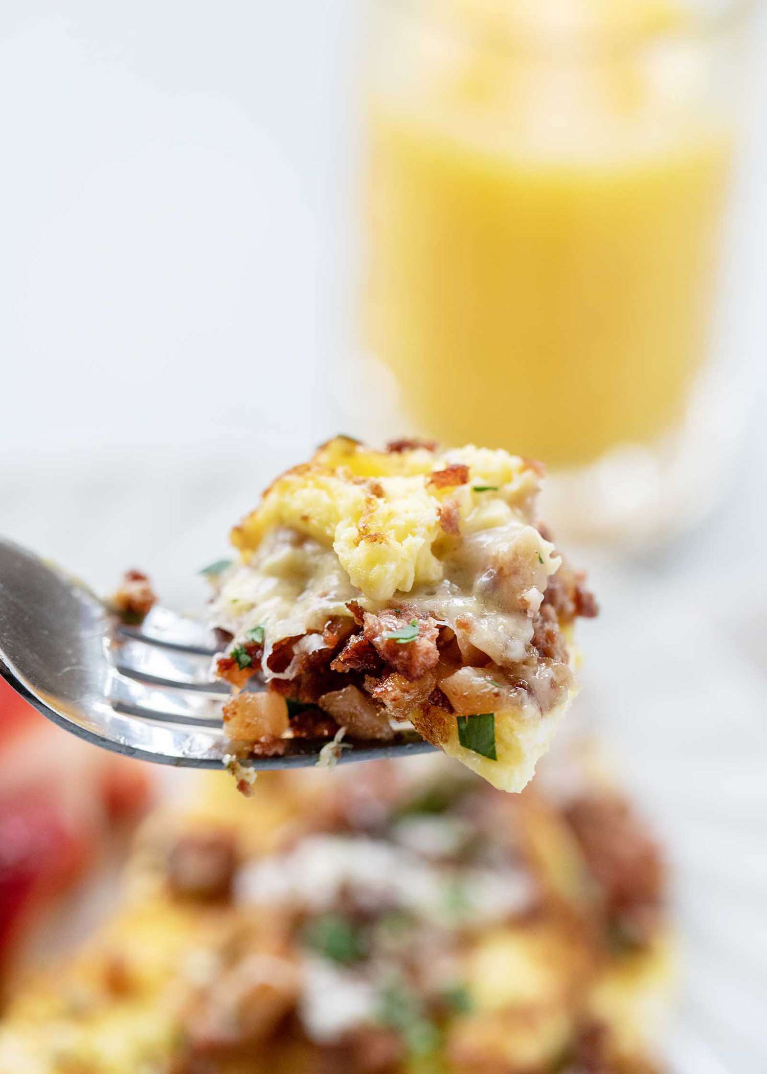 A forkful of the perfect corned beef omelet is in focus. Egg, cheese, corned beef and parsely are visible. The plate of omelet is below the fork. A glass of orange juice is above the fork.