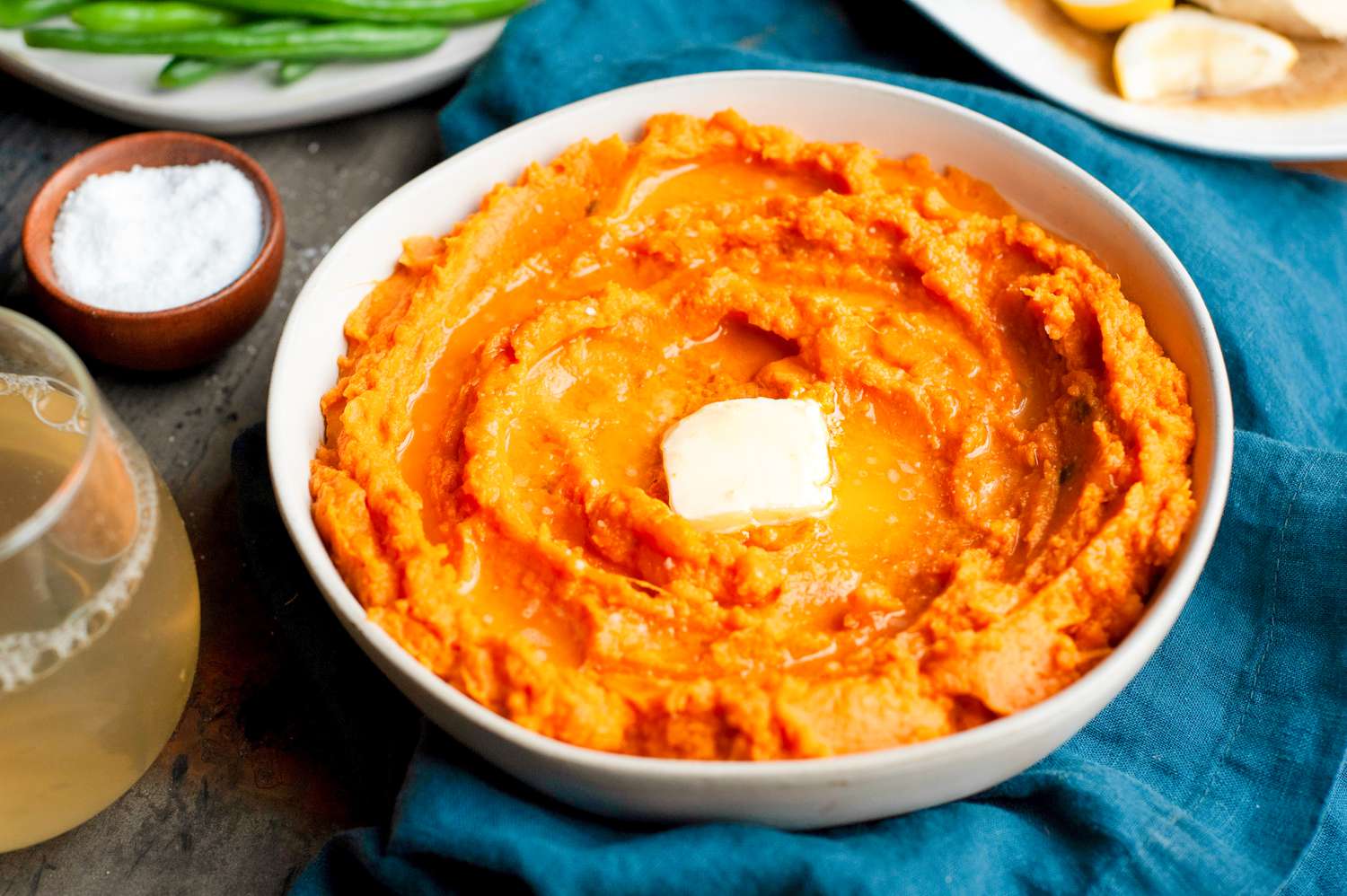 Table Setting with a Bowl of Instant Pot Mashed Potatoes Topped with Butter, a Plate of Green Beans, a Platter of Chicken, a Small Bowl of Salt, a Drink, and a Table Napkin