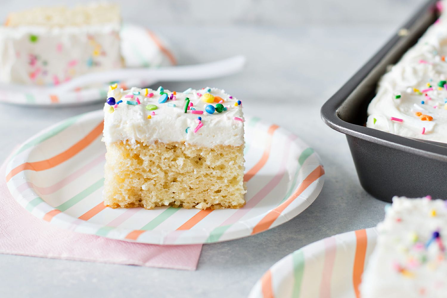 Easy Vanilla Cake Recipe - slice of vanilla cake with white frosting and sprinkles on striped paper plate.