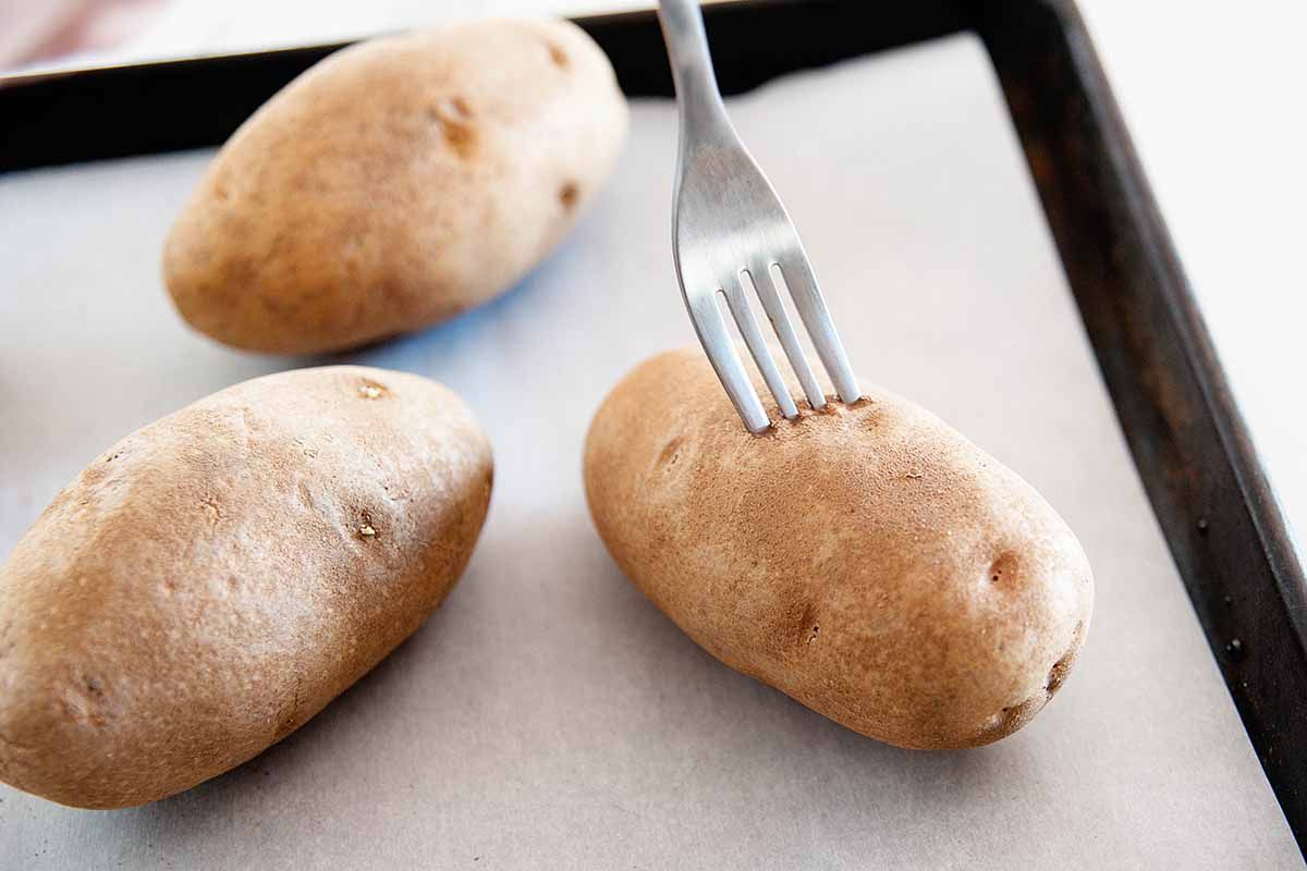 Three potatoes on a baking sheet being priced with a fork