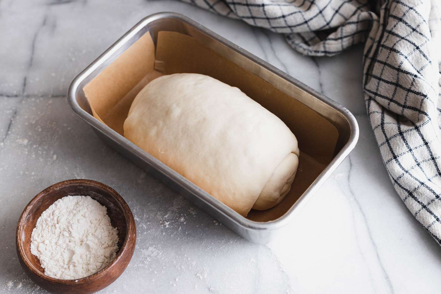 Rolled Sourdough Sandwich Loaf Placed in a Parchment Lined Baking Pan, and Next to It, a Small Bowl of Flour and a Kitchen Towel