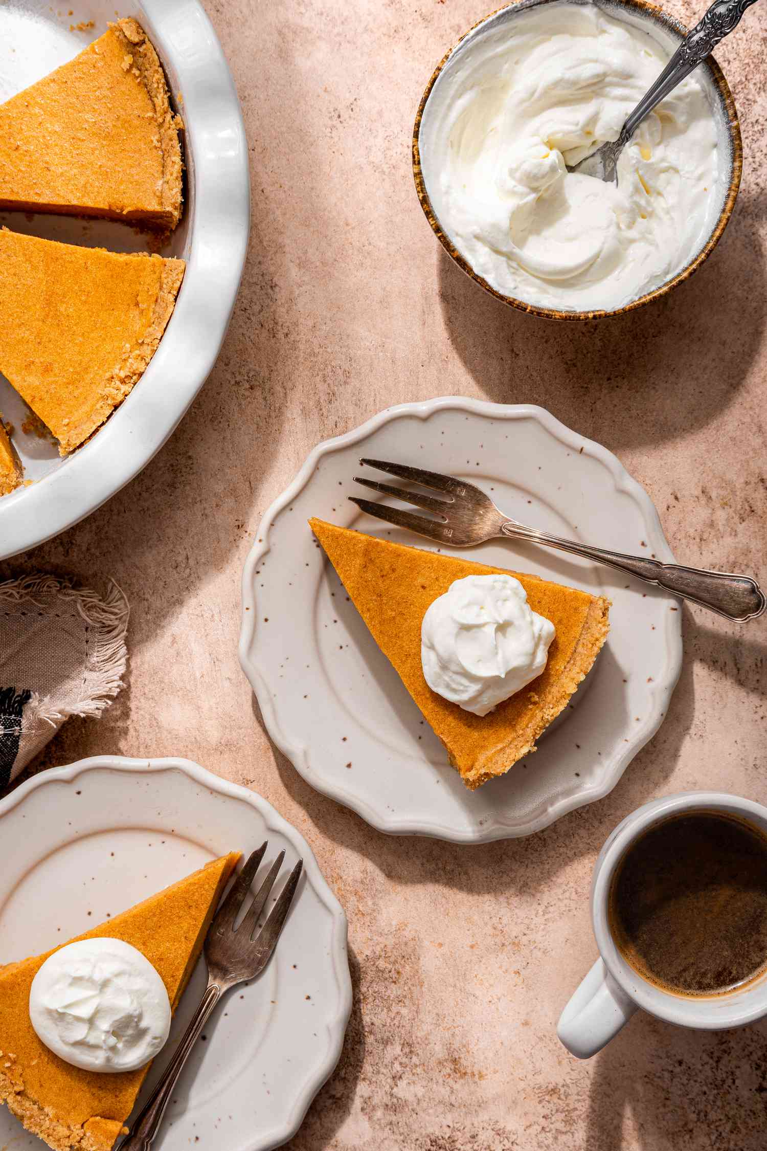 Slices of no-bake pumpkin pie on small plates topped with a dollop of whipped cream and a fork, a bowl of whipped cream, a cup of coffee, and a pumpkin pie in a pie pan