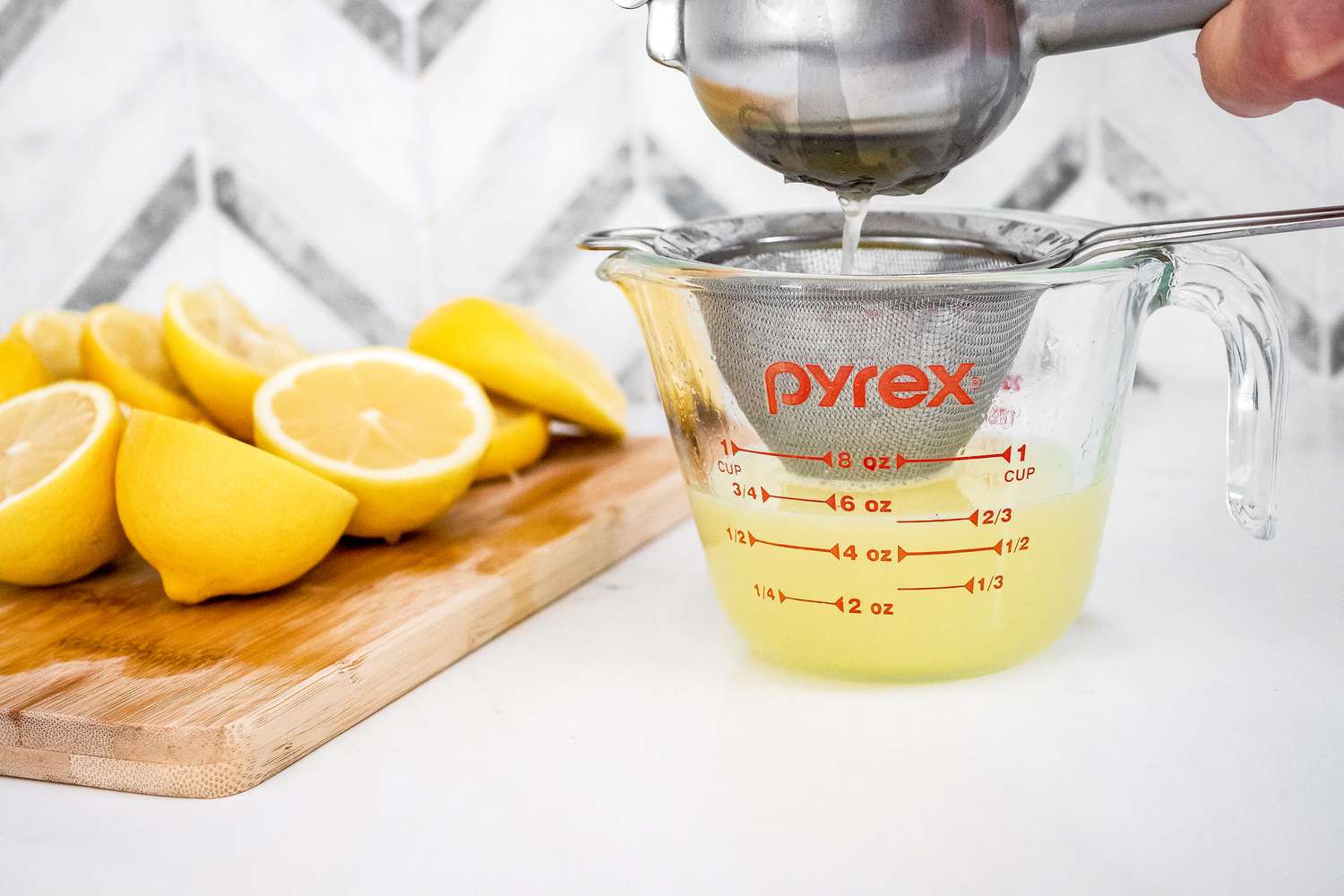 Lemon Pressed Used to Squeeze Lemon Halves Over a Strainer and a Pyrex Measuring Cup, and Next to It, a Cutting Board With Lemon Wedges 