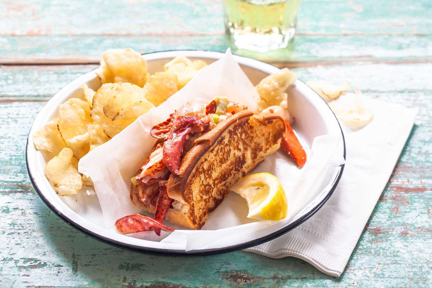 Plate of the best lobster roll recipe served with a side of chips and a lemon wedge.