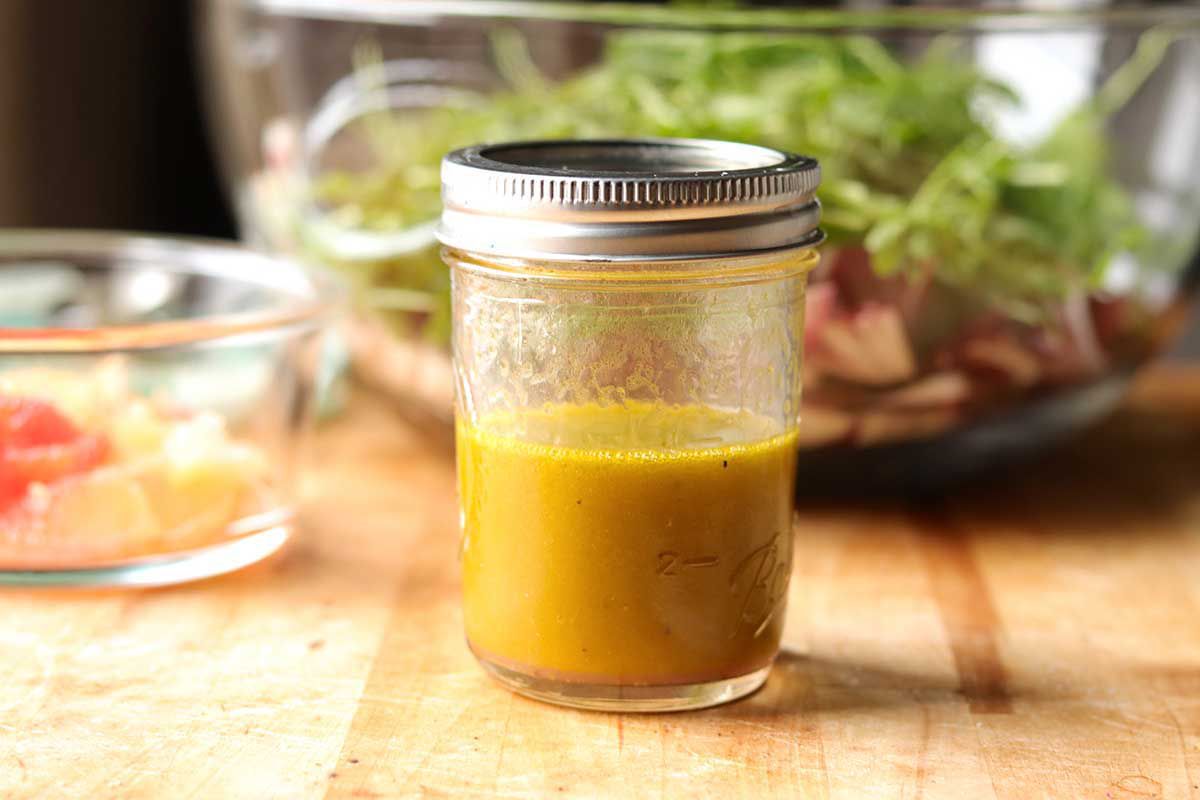A mason jar half full of vinaigrette for a winter salad. The mason jar is on a wooden cutting board and a glass bowl with a few citrus slices is to the left and a large glass bowl with arugula and radicchio is behind the mason jar.
