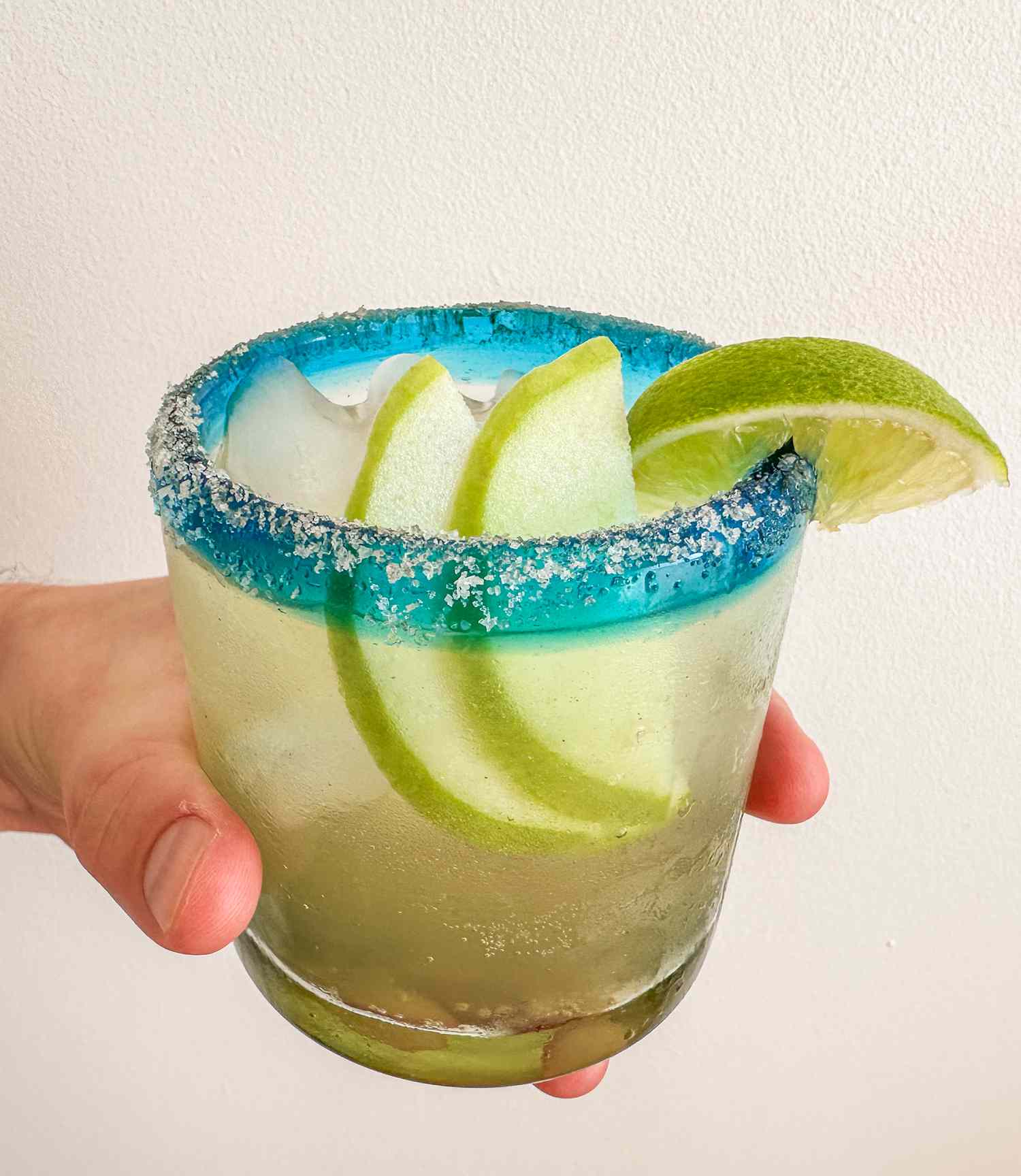 A hand holding a glass of 3-Ingredient Sparkling Apple Margarita with slices of apple inside and salt and lime on the rim against an off-white background
