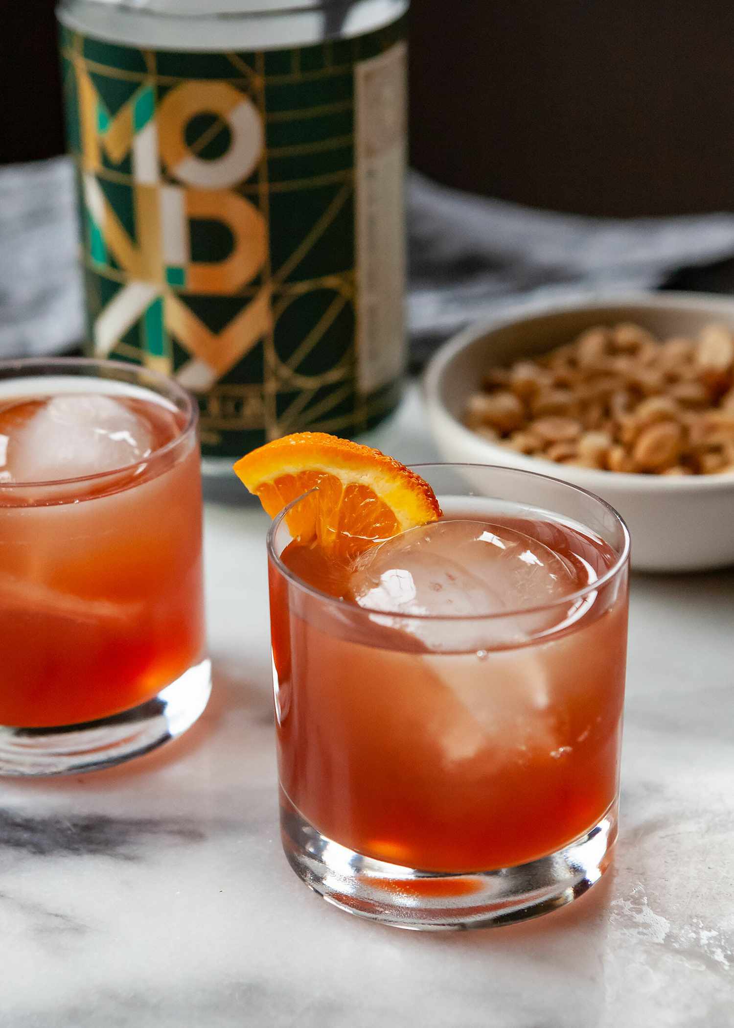 Non alcoholic negroni in a rocks glass with ice and an orange wedge. Bar nuts, and the non alcoholic gin are pictured behind the glass.