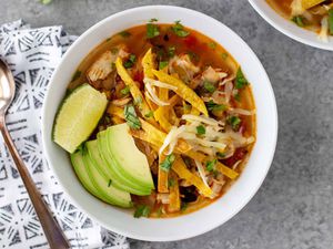 A white bowl of pressure cooker tortilla soup with a spoon and grey patterned linen napkin to the left. The top right corner has a partial view of a second bowl of the same soup.