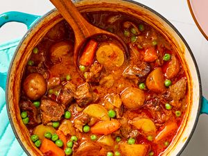 Overhead closeup view of hands-off beef stew in a teal colored dutch oven with a wooden spoon, all on a tile countertop next to a pink bowl and blue hotpad