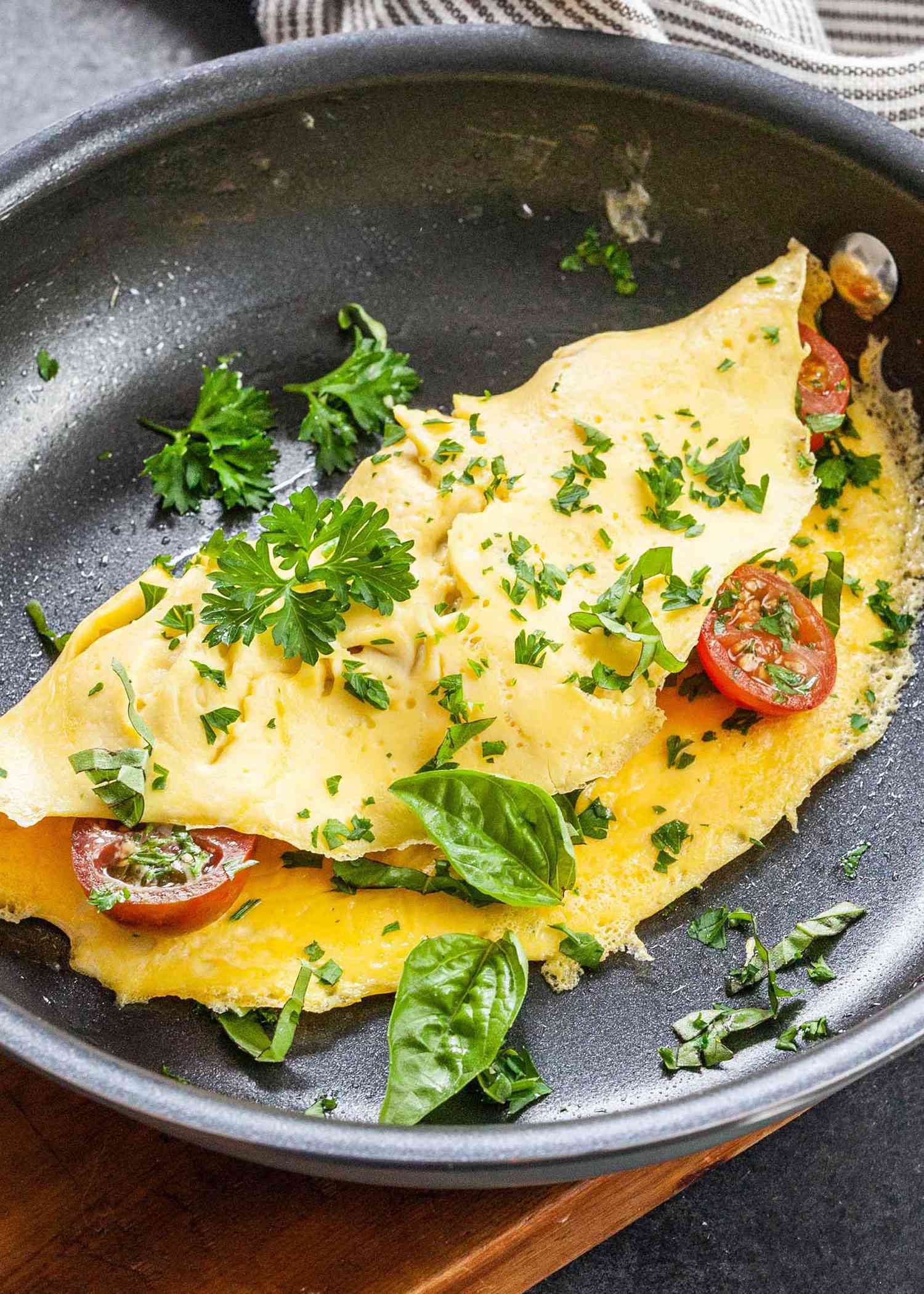 An omelette cooking on a non-stick pan with cut cherry tomatoes inside and parsley sprinkled on top