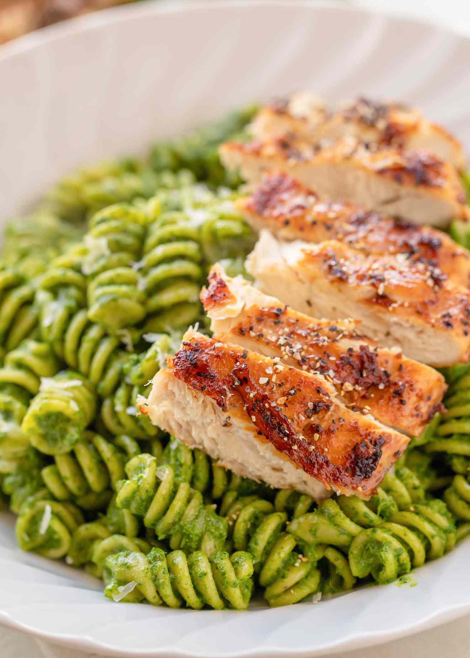 Close up view of easy rotini kale pesto in a white bowl. Roasted chicken breast is sliced and laid on the right side.