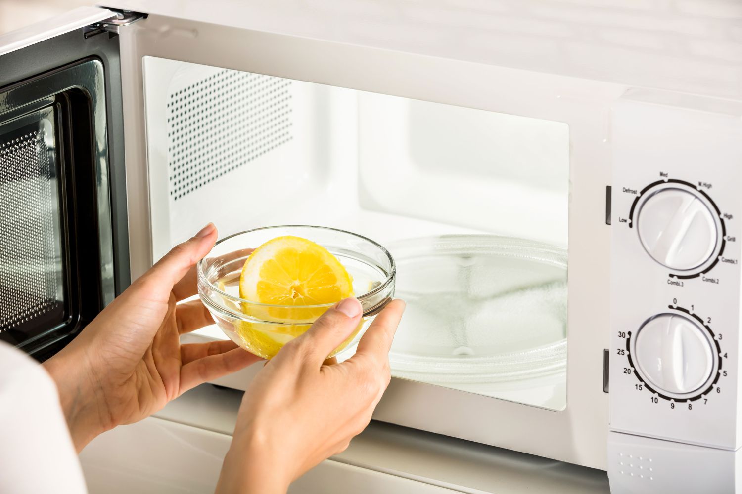 Person placing a glass bowl with a lemon slice into a microwave