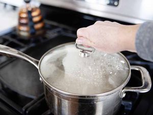 Silver saucepan with a hand grasping the lid handle. Pan sits on gas stove.
