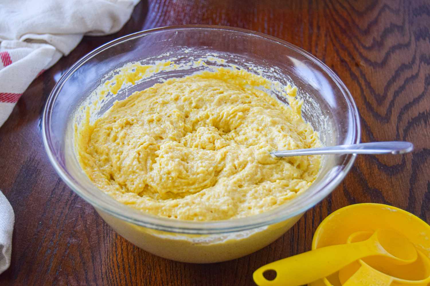 Cornbread batter and a spoon in a glass bowl with measuring cups on the side