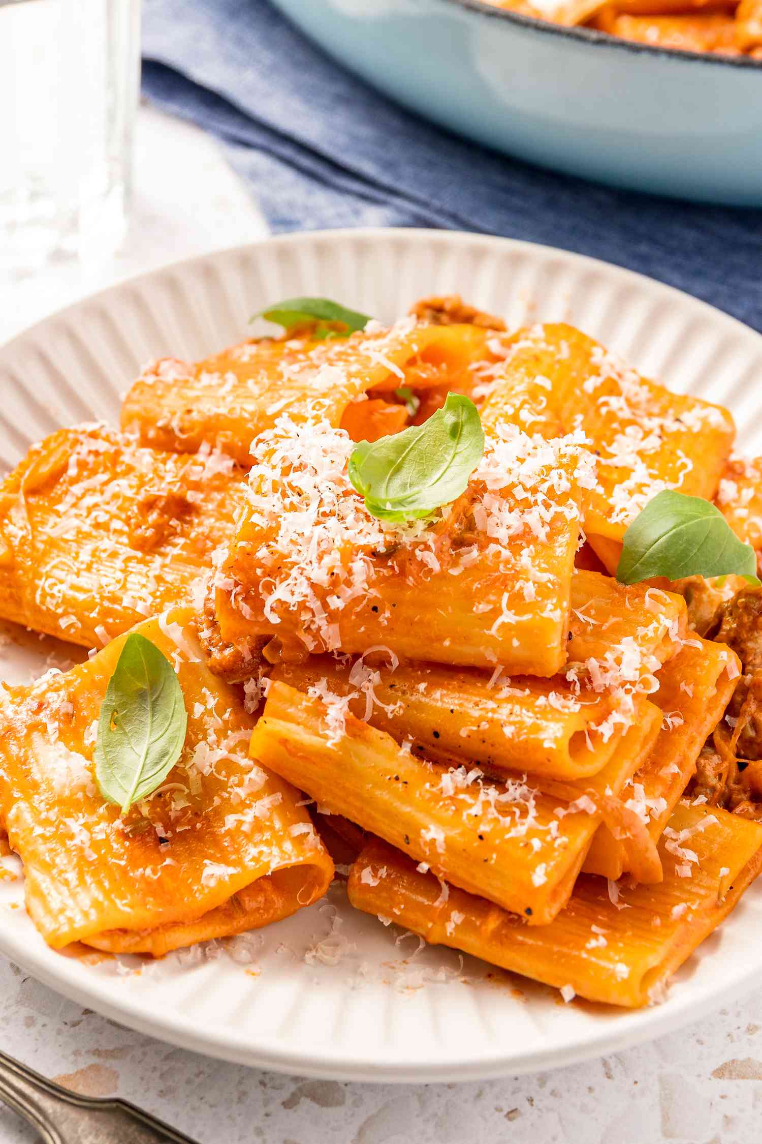 Plate of Paccheri With Quick Sausage Ragu Topped With Basil Leaves, and in the Surroundings, a Glass of Water and a Pot of More Pasta