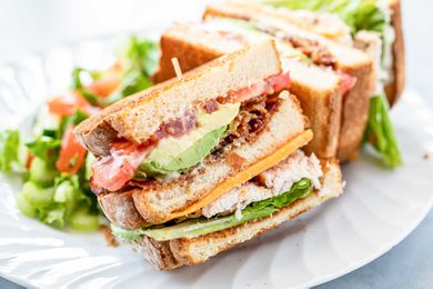 Side view of turkey club with garlic mayo on a plate. The bread is toasted and the mayo, avocado, bacon, tomato, lettuce and turkey are visible in the sandwich.