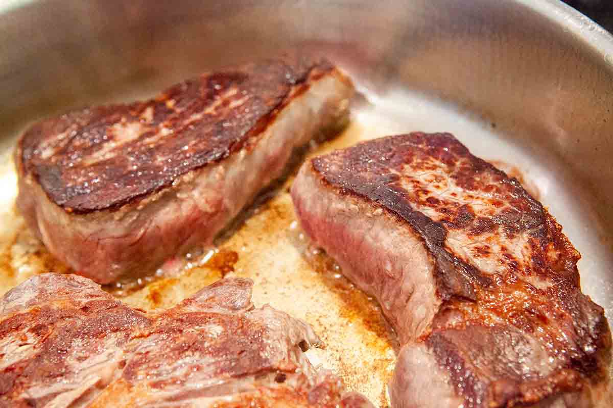 steaks searing in a pan for peppercorn steaks