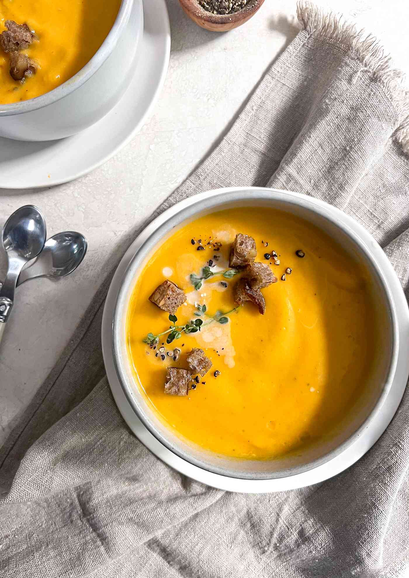 Bowl of Vegan Butternut Squash Soup Topped with Thyme, Coconut Milk, Croutons, and Cracked Pepper, Sitting on Top of a Kitchen Linen and Next to Two Spoons and Another Bowl of Soup