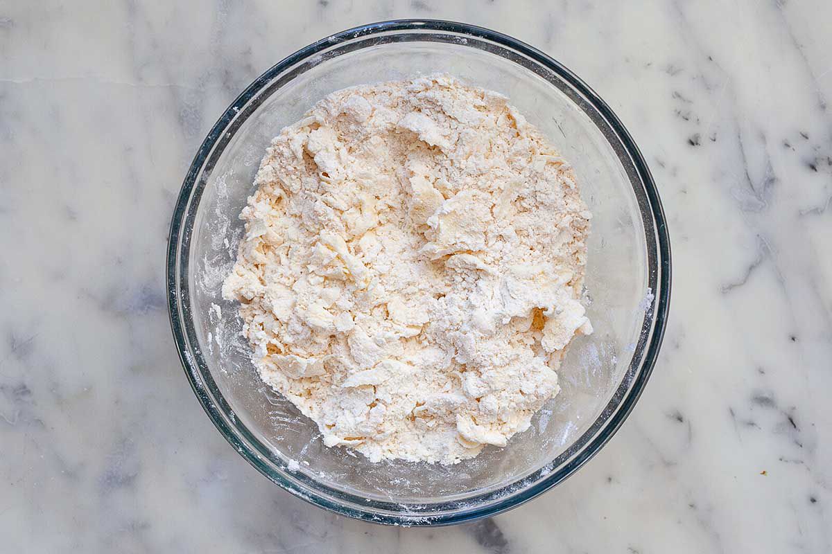 A glass bowl is on a marble countertop and the dough mixture for the homemade quiche crust is inside.