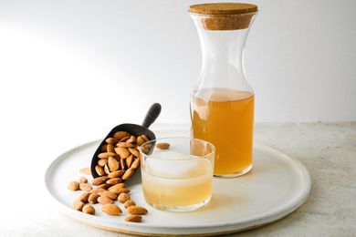 Homemade orgeat in a pitcher behind a cocktail and a scoop with almonds.