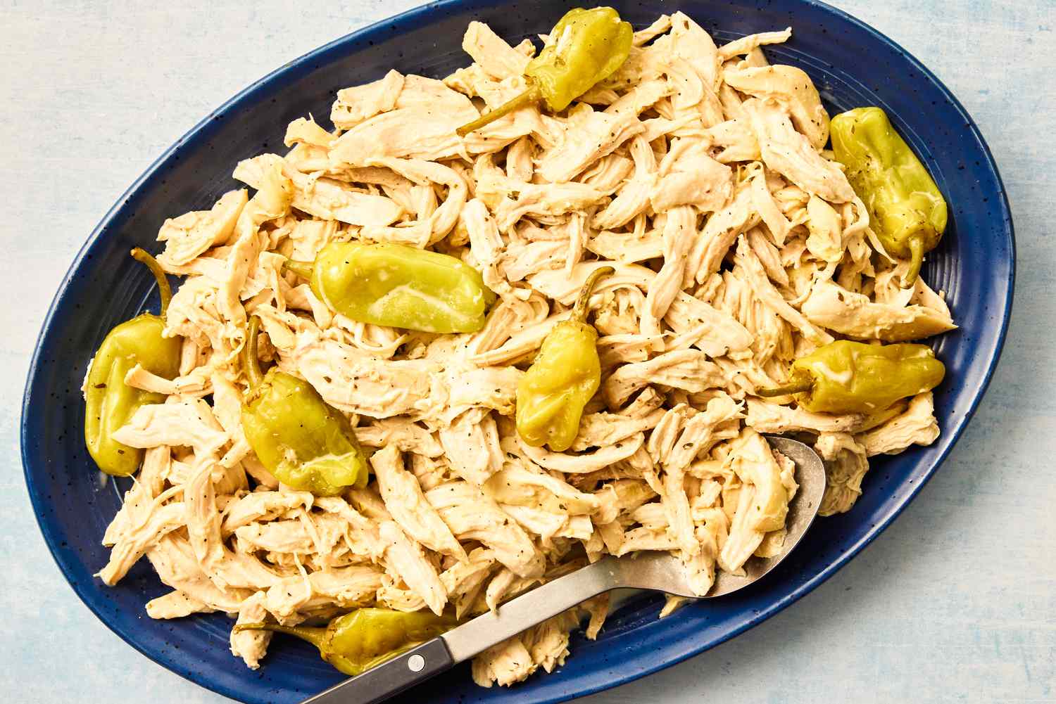 Overhead view of a blue platter and a serving spoon with shredded chicken and pepperoncini