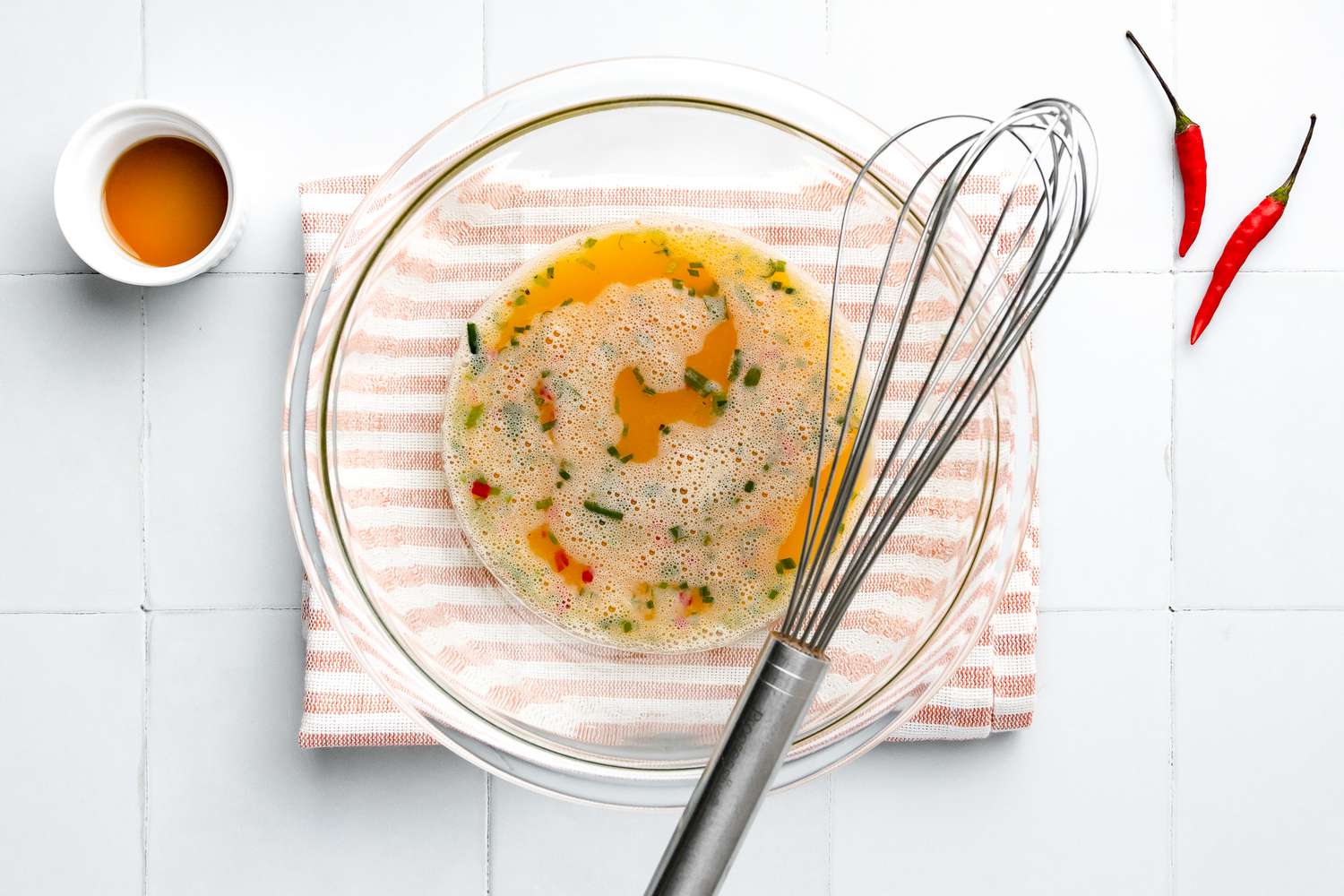 Bowl of Whisked Kai Jeow Ingredients with Bowl Sitting on a Kitchen Towel and Next to It, a Small Bowl of Fish Oil and Two Thai Chilis