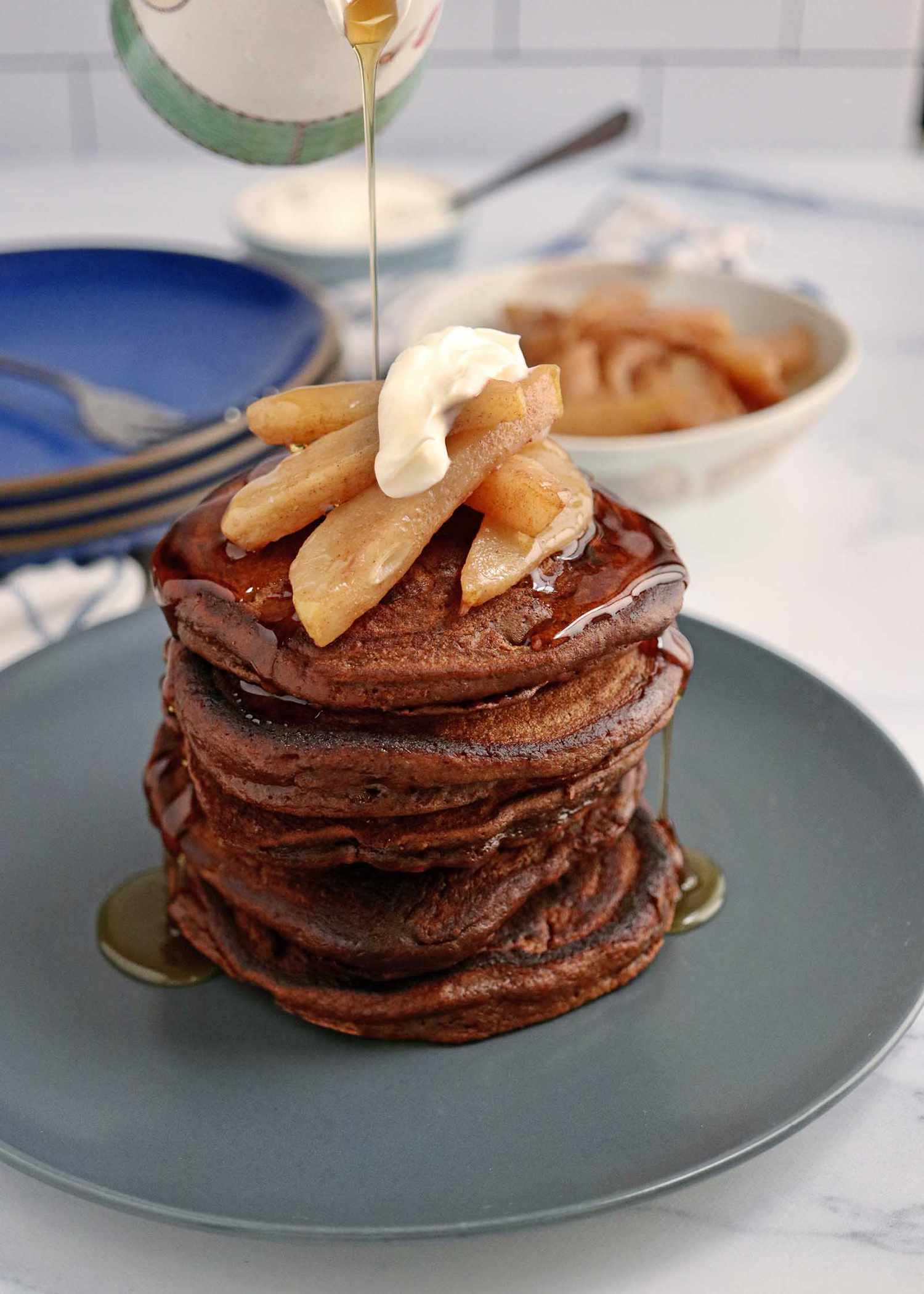 A stack of holiday pancakes topped with sliced pears and creme fraiche with syrup being poured over top.