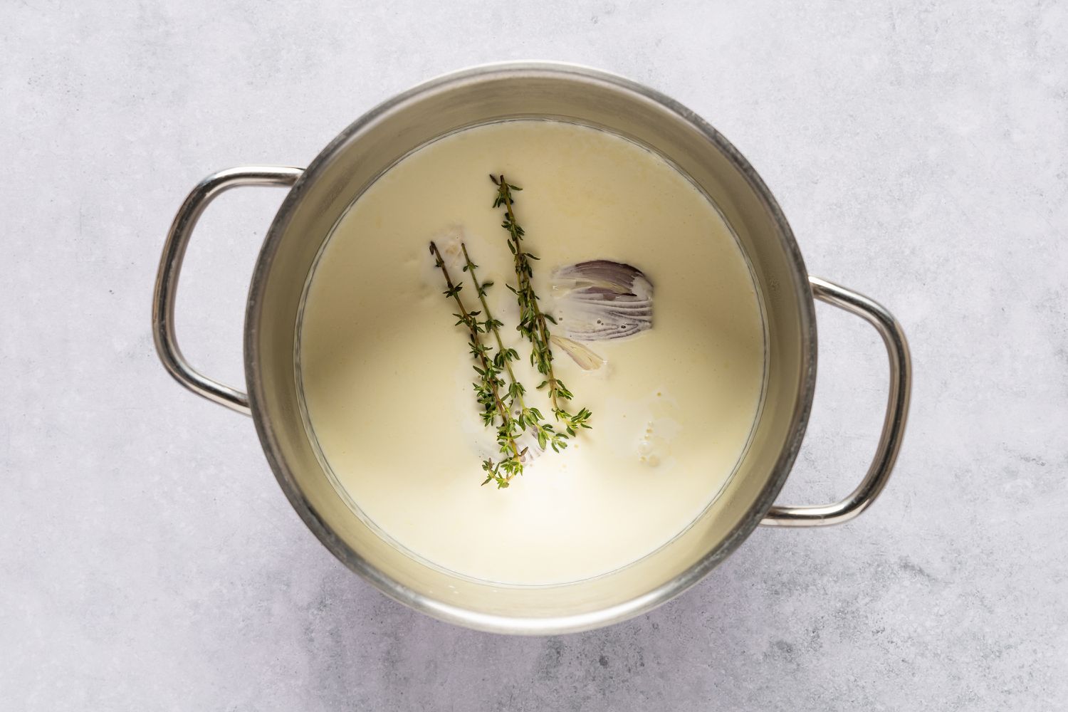 Overhead view of a small pot of the cream with sprigs of thyme, shallot halves and garlic cloves for Sheet Pan Au Gratin Potatoes recipe