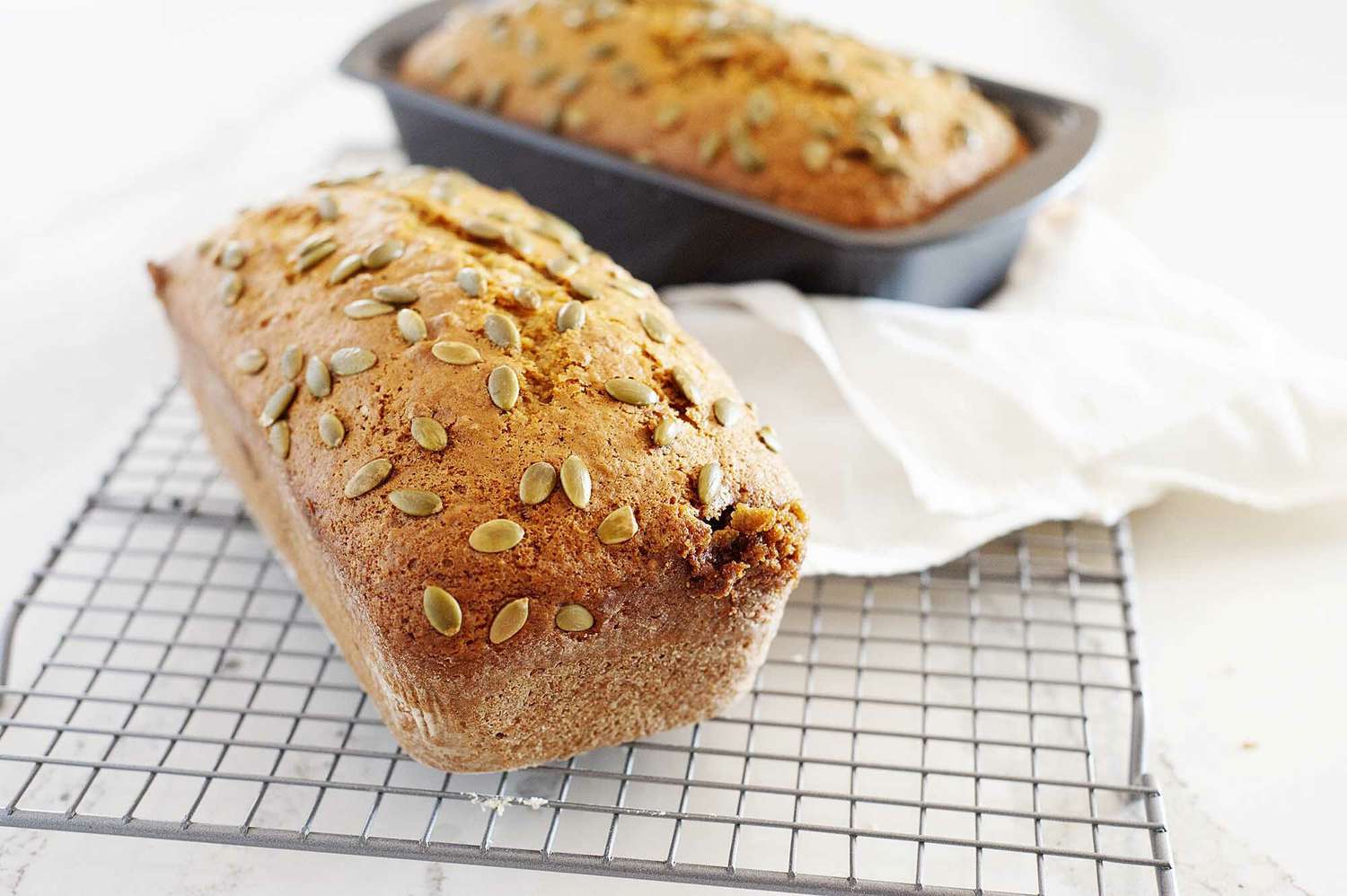 A loaf of Moist Pumpkin Bread cooling on a rack with a second loaf in the pan behind it.