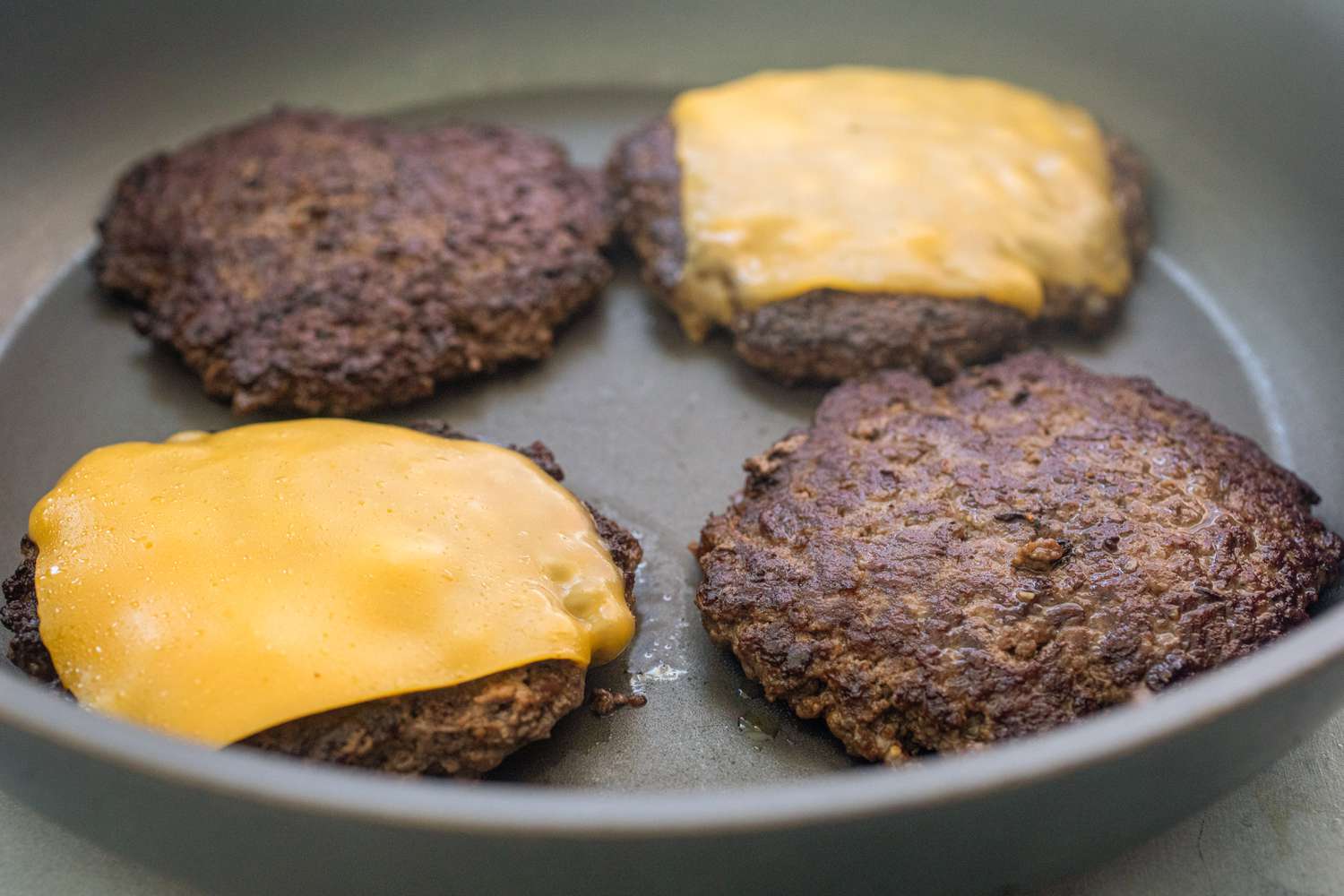 Four burger patties in a skillet with two topped with cheese to show how to make smashburgers