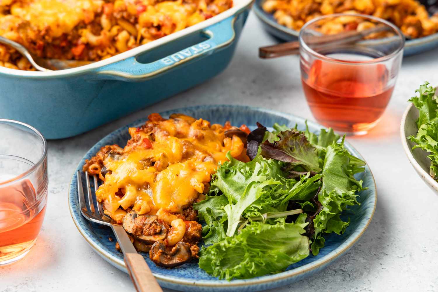 Serving of Johnny Marzetti casserole and mixed greens on a plate at a table setting with more casserole in a casserole dish, another plate with another serving and two glasses with a drink