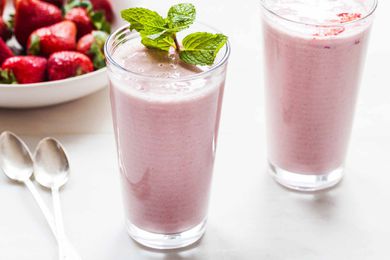 Two tall glasses of healthy strawberry almond oat smoothies on a white counter. A sprig of mint is on the top of one glass two spoons and a bowl of strawberries are to the left of the glasses.