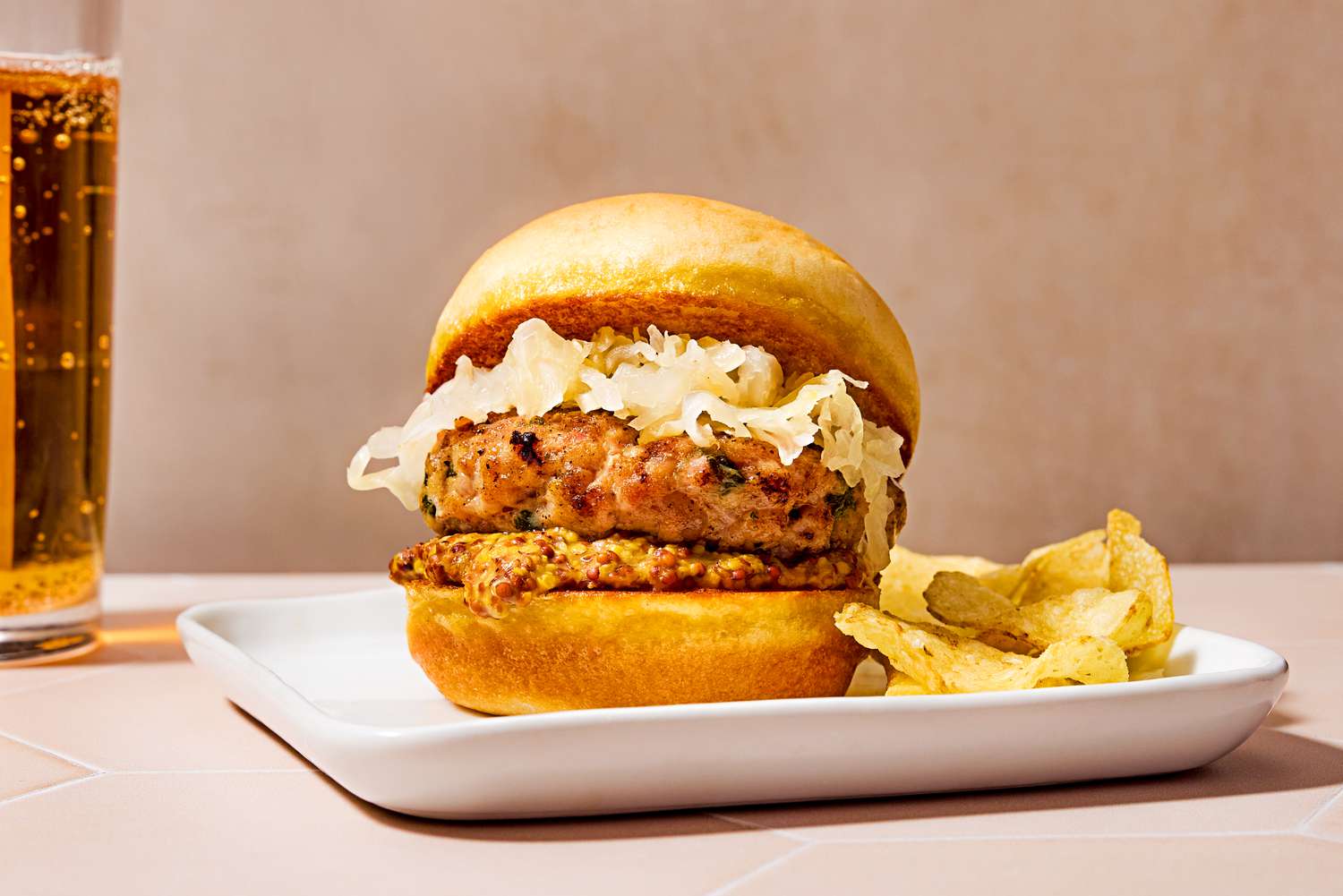 Side view of a bratwurst slider topped with sauerkraut, on a plate with a side of potato chips and a glass of beer in the background