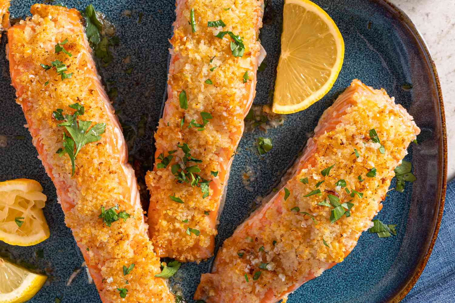 Overhead view of a blue plate of three salmon fillets topped with garlic butter, panko, and parmesan mixture and sprinkled with parsley next to several lemon slices