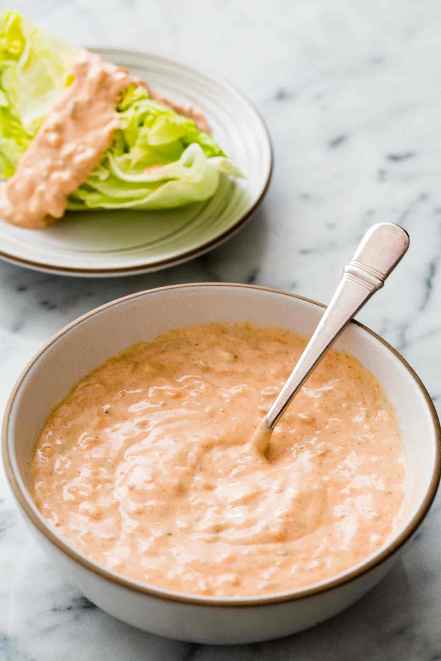 A bowl of thousand island dressing with a plate of salad dressed already in the background