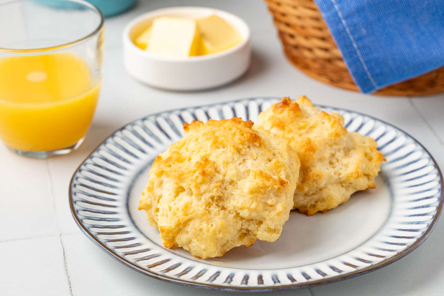 Drop biscuit on a small plate, and in the surroundings, a glass of orange juice, a small bowl of butter, and the basket with more biscuits